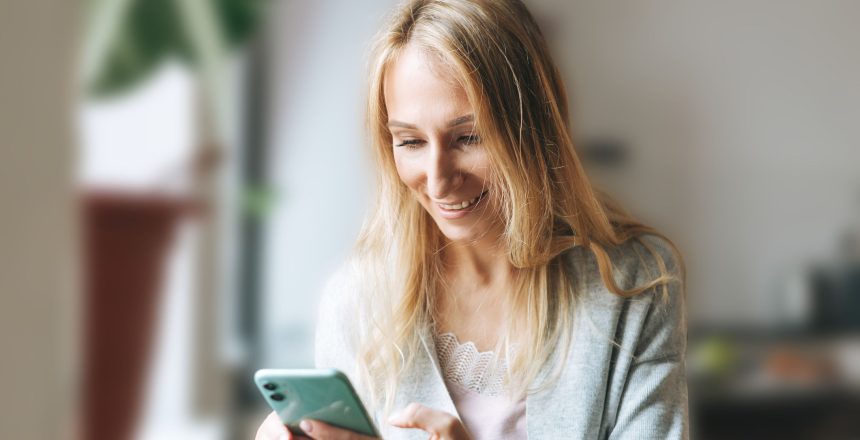 Beautiful blonde young woman in home wear using mobile in kitchen at home